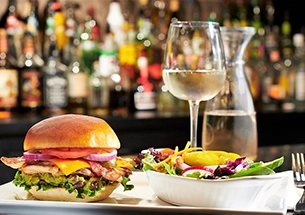 Lunch menu item featuring a gourmet grilled chicken burger with cheese and bacon, served alongside a fresh side salad and a glass of white wine at Symposium Cafe Restaurant in Bolton